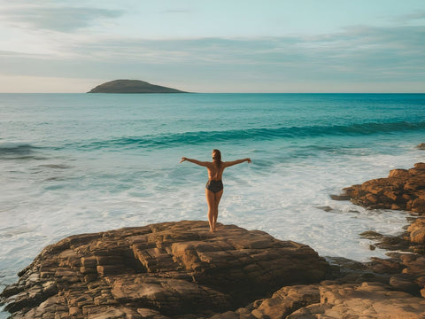 A woman standing on coastal rocks looking out over the ocean on a summer morning