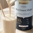 A thick creamy vanilla Slim Down Shake being poured into a glass, beside the 840g Creamy Vanilla tin showing 30g protein and 7g fibre per serve.