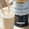 A thick creamy vanilla Slim Down Shake being poured into a glass, beside the 840g Creamy Vanilla tin showing 30g protein and 7g fibre per serve.
