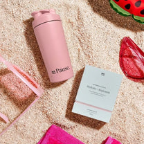 Pink MyPause steel shaker bottle and a Hydrate + Replenish box on sand, with sunglasses and a watermelon slice.